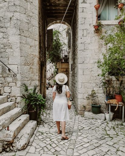 Rear view of young woman wearing white dress and sun hat holding handbag. Lifestyle and fashion full length image of female tourist in old town.