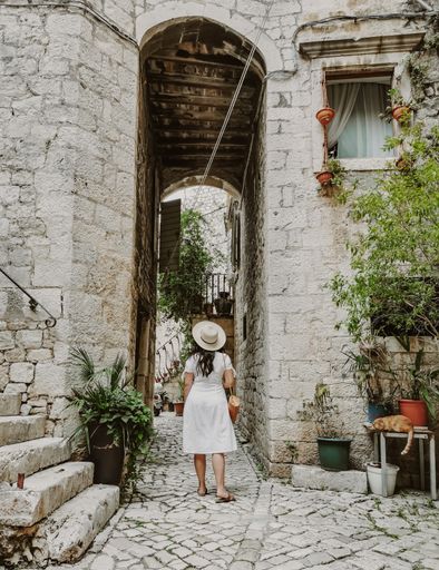 Rear view of young woman wearing white dress and sun hat holding handbag. Lifestyle and fashion full length image of female tourist in old town.