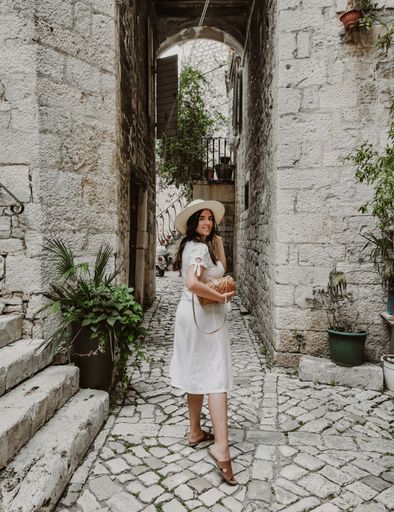 Side view of young woman wearing white dress and sun hat holding handbag. Lifestyle and fashion full length image of female tourist in old town.