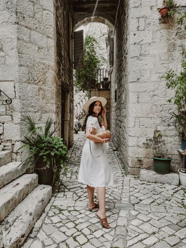 Side view of young woman wearing white dress and sun hat holding handbag. Lifestyle and fashion full length image of female tourist in old town.