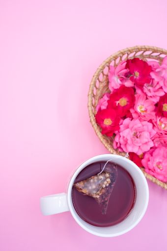 A vertical top view of a basket full of red and pink flowers next to a cup of tea with copy space above on a vibrant pink background