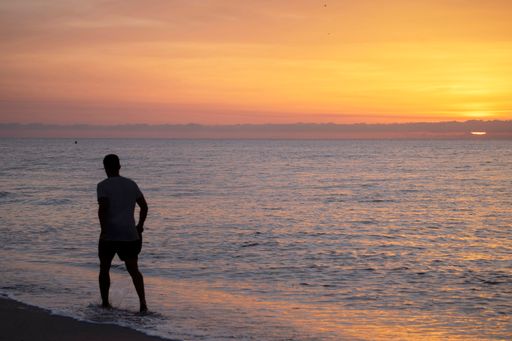 A man is walking along the sandy beach at sunrise while looking at the ocean. The sky is a beautiful mix of orange and pink tones. Beach vacation