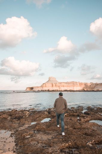 A man strolling on a rocky shore by the ocean under a cloudy blue sky