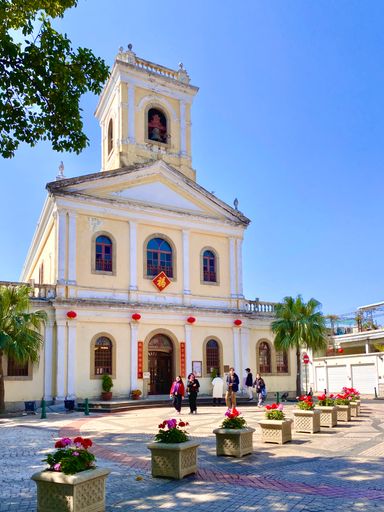 Visitors near a Portuguese-style church with bell tower in Macao, surrounded by palm trees and red lanterns.