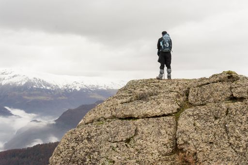 A back view of a hiker with a backpack standing on a rock admiring the snow-capped mountains in winter