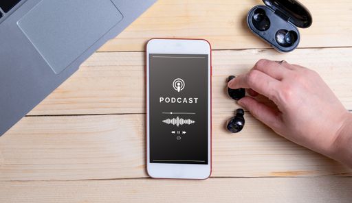 A top view of a smartphone playing a podcast and a hand connecting wireless earphones on a wooden table