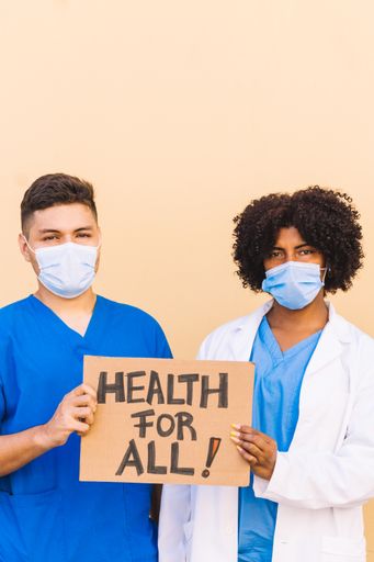 A vertical shot of Hispanic and Afro-American doctors or nurses wearing uniforms and sanitary masks holding a "Health for all" banner