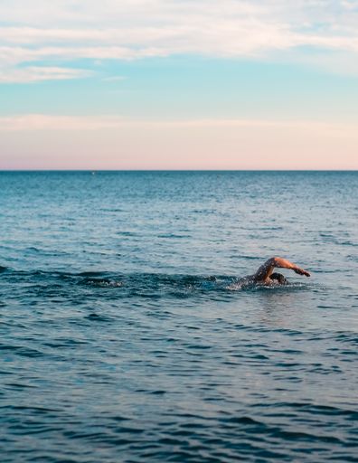 A vertical shot of a man swimming in a beautiful blue ocean with small waves and a bright clear sky