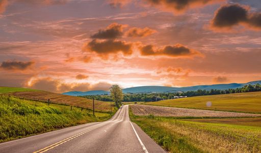 A long straight empty road through the green grassy field under bright cloudy sunset sky