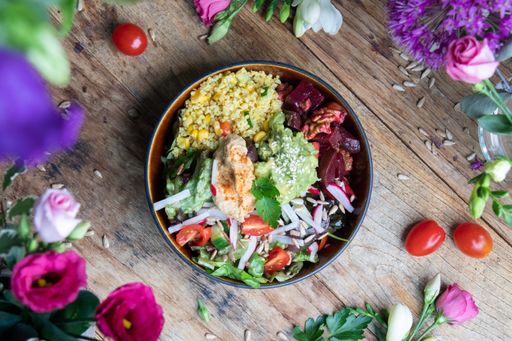 A top view of a bowl of healthy salad with avocado, vegetables, and seeds on a wooden table among flowers