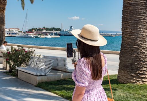 Female tourist walking on seaside promenade in Split, Croatia. Young woman using mobile phone and gimbal to take photos and record video.