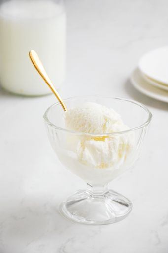 A vertical shot of scoop of vanilla ice cream in a glass bowl with a golden spoon on a white background