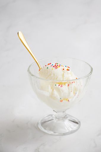 A vertical shot of bowl of vanilla ice cream with colorful sprinkles and a golden spoon on a white background
