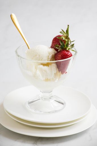A vertical shot of glass bowl of vanilla ice cream topped with fresh strawberries and a golden spoon on white plates