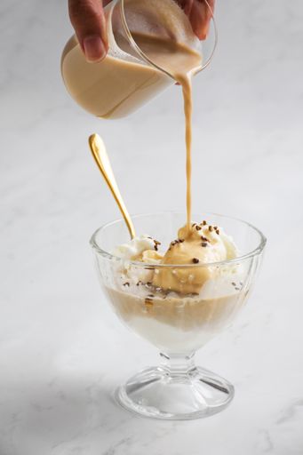 A vertical shot of a hand pouring coffee over ice cream in a glass cup, creating an affogato dessert with a golden spoon