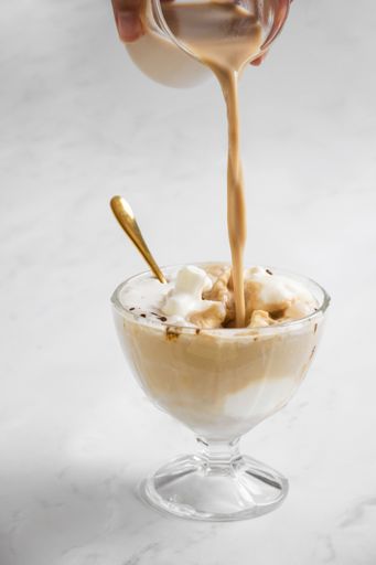 A close-up vertical shot of a creamy dessert being poured into a glass bowl with a golden spoon on a white background