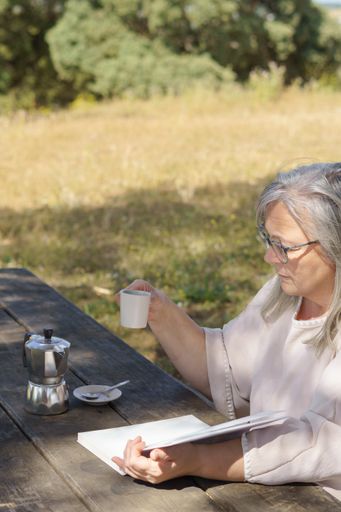 woman reads a book at a table in the field while drinking a cup of coffee in the background of a grassy meadow.
