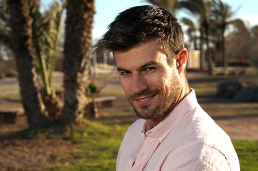 A closeup shot of a handsome male looking invitingly at the camera with a one-sided smile and a mild pink classic shirt