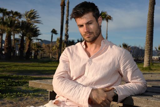 A young Hispanic male wearing a pink shirt and posing on the beach near palm trees