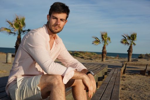 A young Hispanic male wearing a pink shirt and posing on the beach near palm trees