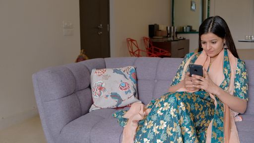 A young South Asian woman in traditional clothing sitting on a purple couch texting on her phone