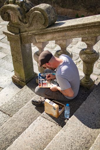 A Man sitting on old stairs with a box and painting on a board on his lap, wearing a beret