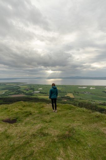 A vertical shot of a caucasian female with a blue jacket standing in a green field with a view of the sea