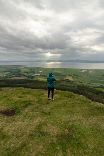 A vertical shot of a caucasian female with a blue jacket standing in a green field with a view of the sea