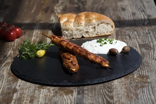 A closeup shot of a minced meat skewer with white sauce and a piece of bread on a rustic table