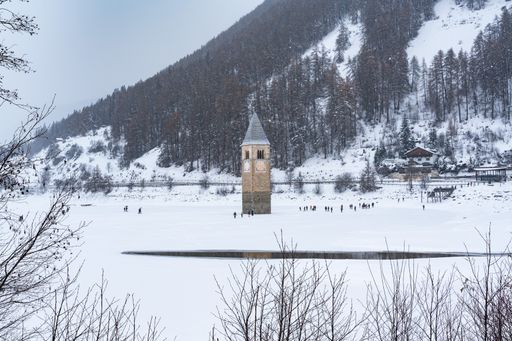 A bell tower stands prominently in the center of snow-covered Resia Lake, surrounded by a serene winter landscape with people walking around
