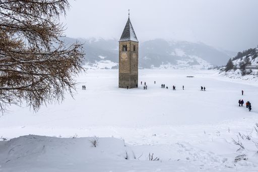 The bell tower stands prominently in the center of snow-covered Resia Lake, surrounded by a serene winter landscape, as people wlak around it