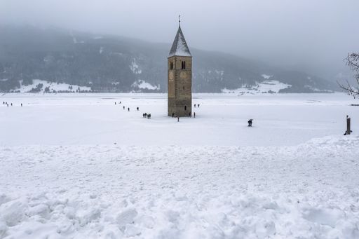 A bell tower standing in the middle of Resia Lake during winter with people walking around it