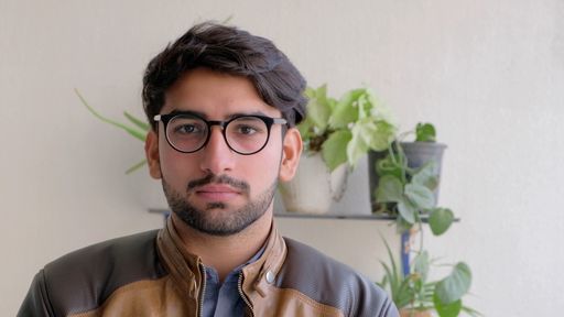 A young Indian male in eyeglasses wearing a brown leather jacket with houseplants in the background