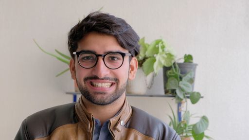 A happy young Indian male in eyeglasses wearing a leather jacket with houseplants in the background