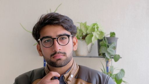 A view of an Indian guy with glasses and a leather jacket standing in front of plants at home