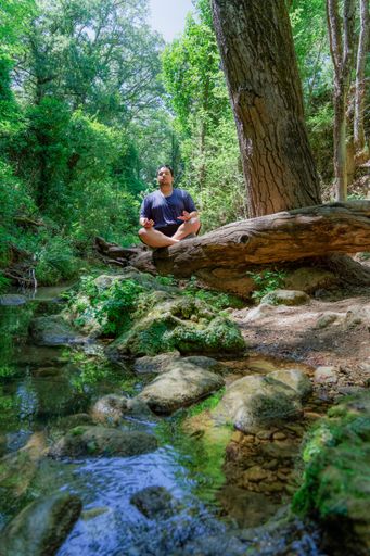 young dark-haired boy in a blue T-shirt meditating sitting on a tree trunk over a river in a tropical rainforest