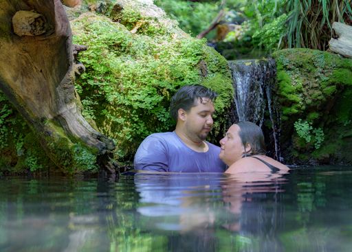 mother and son embrace looking at each other while bathing in a river under a mossy waterfall