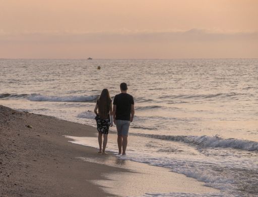A couple holding hands while walking on the fine sand of the brown beach by the beach in the warm sunrise.