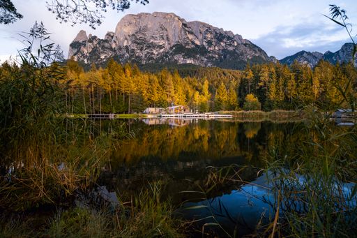 A solitary house with a prominent mountain backdrop, shot during sunset golden hour with a lake in the foreground reflecting the forests