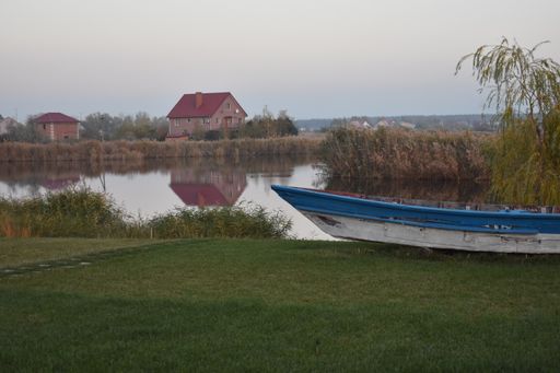 A closeup of a thin boat parked on vibrant green grass with a lake and an old house in the background