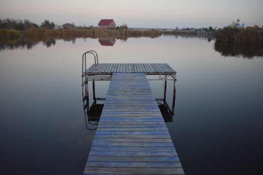 A rusted wooden dock on a clear blue lake with grass on the sides reflecting in it on a chilly day