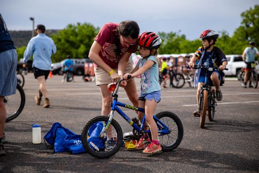 A mother teaching her daughter to ride a bike in a parking lot with other cyclists in the background