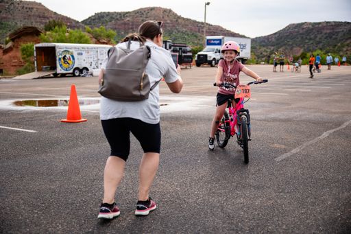 A young girl riding a bicycle with a helmet on in a parking lot while an adult takes a photo