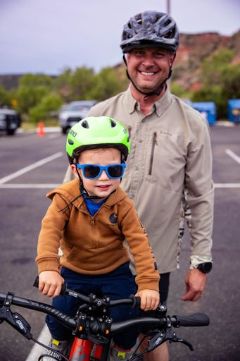 A man and a young boy, both wearing helmets, enjoying a bike ride in an otdoor parking area.