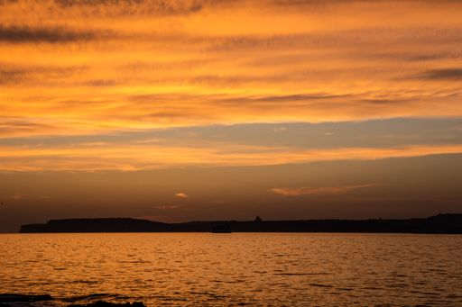 Sun below horizon, stratus, cirrus clouds lit by orange afterglow;ferry at sea; Gozo, church dome silhouetted in background, Gozo-Malta Channel, Malta