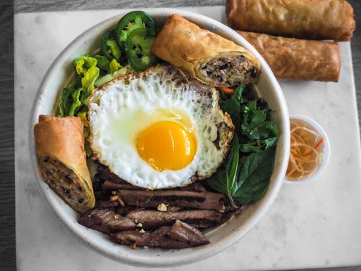 An overhead shot of a dish with fried eggs and meat with vegetables and spring rolls on top