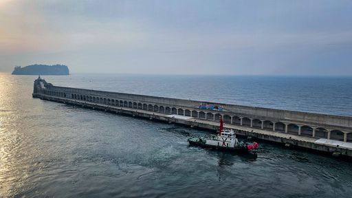 A serene view of a pier extending into the sea with a small tugboat and an island in the distance during sunset.