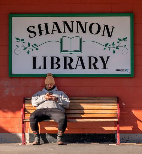 A young person sitting on a bench outside Shannon Library, wearing a beanie and using a smartphone