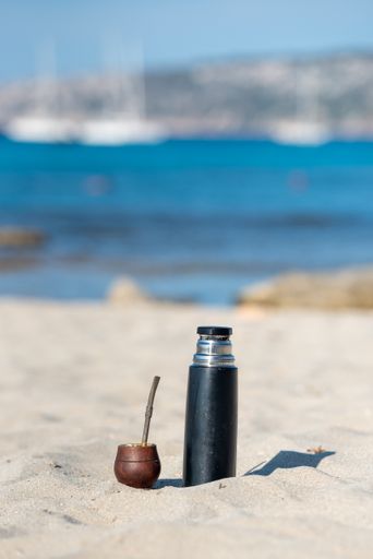 A vertical shot of a cup of mate tea with a straw and a thermos flask on the beach on a sunny da