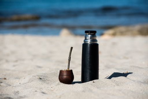 A closeup of a cup of mate tea with a straw and a thermos flask on the beach on a sunny day